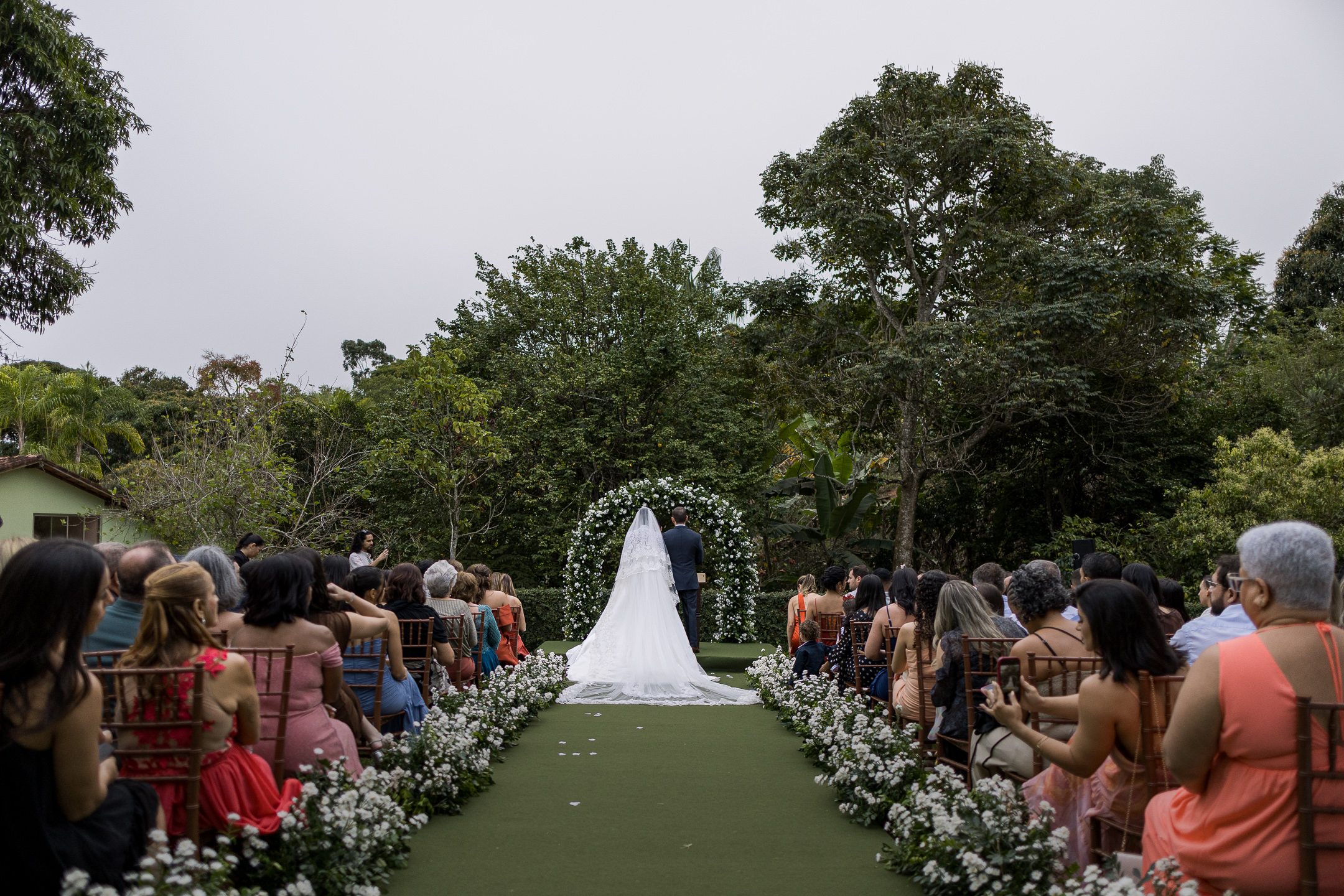 Corredor da cerimônia nos jardins, ladeado de flores brancas, com o arco florido ao fundo